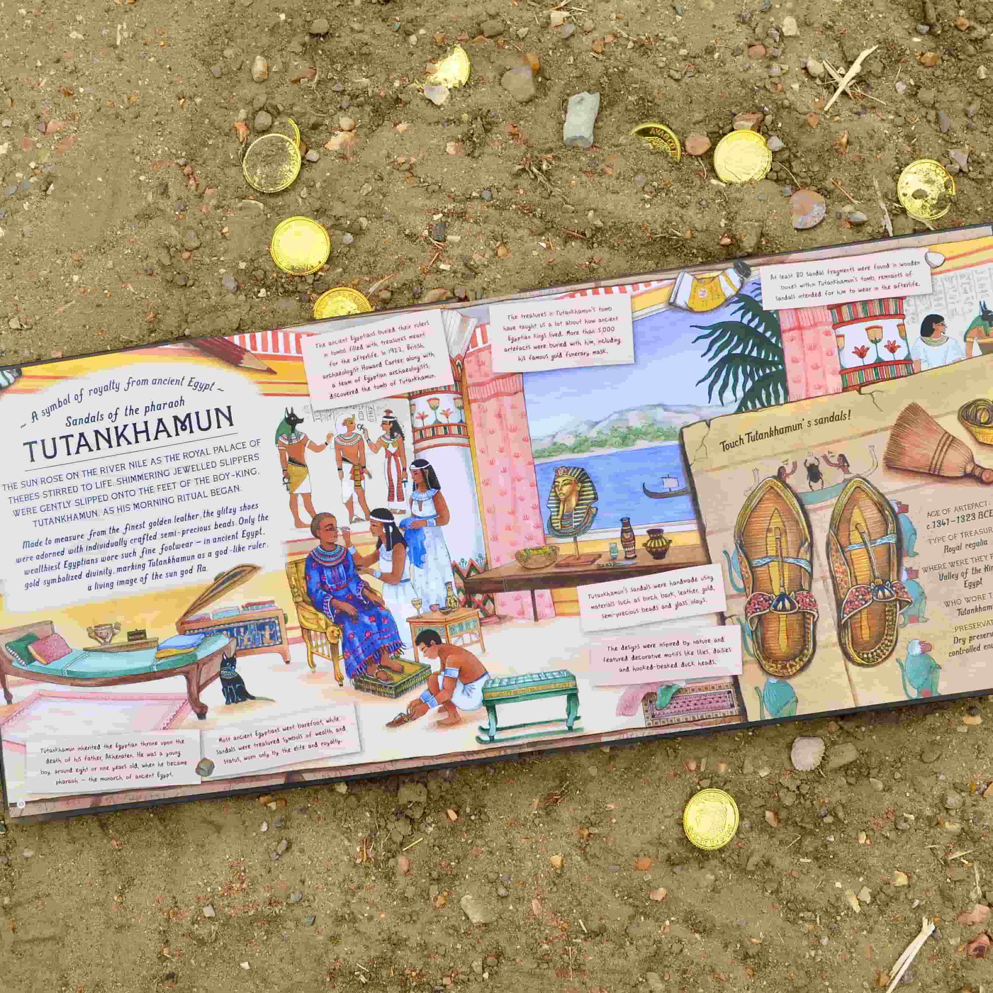 A children’s book page about Tutankhamun lies open on sandy ground with scattered gold coins, featuring colourful Egyptian illustrations and fascinating archaeological discoveries about the pharaoh.